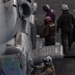 Military personnel are conducting pre-flight or post-flight operations on a naval flight deck alongside a military helicopter, likely a CH-53 or similar heavy-lift rotary-wing aircraft. At least four individuals are visible, wearing different colored helmets and flight gear indicating various crew roles. The scene appears to take place on the deck of a naval vessel.