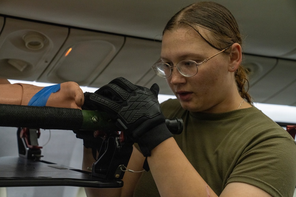 A young person wearing an olive green t-shirt, round wire-framed glasses, and black tactical gloves is focused on assembling or adjusting a piece of equipment, possibly a drone or mechanical device. The setting appears to be an indoor workspace with fluorescent lighting and a low ceiling. The individual has reddish-brown hair pulled back in a braid and is wearing a silver bracelet.