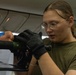 A young person wearing an olive green t-shirt, round wire-framed glasses, and black tactical gloves is focused on assembling or adjusting a piece of equipment, possibly a drone or mechanical device. The setting appears to be an indoor workspace with fluorescent lighting and a low ceiling. The individual has reddish-brown hair pulled back in a braid and is wearing a silver bracelet.