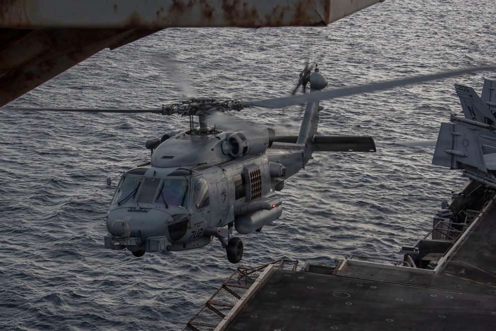 A U.S. Navy MH-60R Seahawk helicopter, marked with the number '702', is hovering or departing from the flight deck of a naval vessel over open ocean. The helicopter is in a gray naval paint scheme and appears to be equipped with sensors and external stores. The image is taken from an elevated position on the ship, likely from a sponson or island structure.