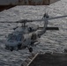 A U.S. Navy MH-60R Seahawk helicopter, marked with the number '702', is hovering or departing from the flight deck of a naval vessel over open ocean. The helicopter is in a gray naval paint scheme and appears to be equipped with sensors and external stores. The image is taken from an elevated position on the ship, likely from a sponson or island structure.