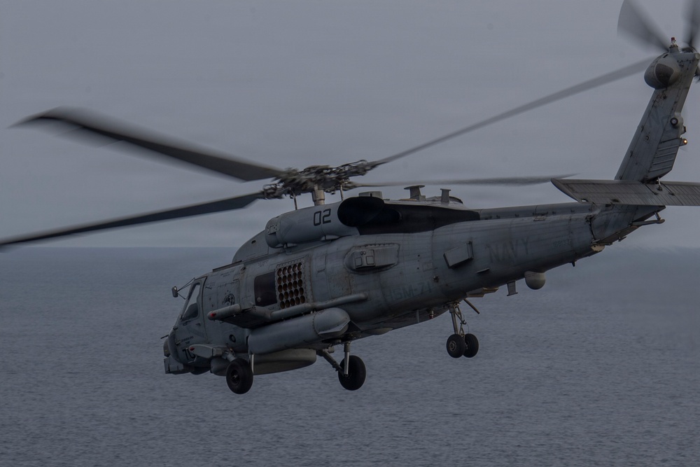 A U.S. Navy MH-60R Seahawk helicopter, marked with the number '02' and 'NAVY' on its fuselage, is captured in low-level flight over open ocean water. The helicopter appears to be in an operational configuration with landing gear deployed and what appear to be weapons or sensor systems mounted externally. The image was taken in overcast, low-light conditions giving it a muted, grey tone.