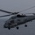 A U.S. Navy MH-60R Seahawk helicopter, marked with the number '02' and 'NAVY' on its fuselage, is captured in low-level flight over open ocean water. The helicopter appears to be in an operational configuration with landing gear deployed and what appear to be weapons or sensor systems mounted externally. The image was taken in overcast, low-light conditions giving it a muted, grey tone.