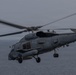 A U.S. Navy MH-60R Seahawk helicopter, marked with the number '02' and 'NAVY' on its fuselage, is captured in low-level flight over open ocean water. The helicopter appears to be in an operational configuration with landing gear deployed and what appear to be weapons or sensor systems mounted externally. The image was taken in overcast, low-light conditions giving it a muted, grey tone.