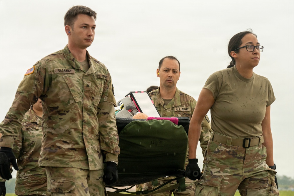 Three U.S. Army soldiers are carrying a litter (stretcher) loaded with gear across an open field under overcast skies. The soldiers are in OCP (Operational Camouflage Pattern) uniforms, with name tapes visible including 'GASAWAY' and partially visible 'KHADKA'. This appears to be a training exercise or field operation involving casualty evacuation or equipment transport.