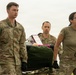 Three U.S. Army soldiers are carrying a litter (stretcher) loaded with gear across an open field under overcast skies. The soldiers are in OCP (Operational Camouflage Pattern) uniforms, with name tapes visible including 'GASAWAY' and partially visible 'KHADKA'. This appears to be a training exercise or field operation involving casualty evacuation or equipment transport.