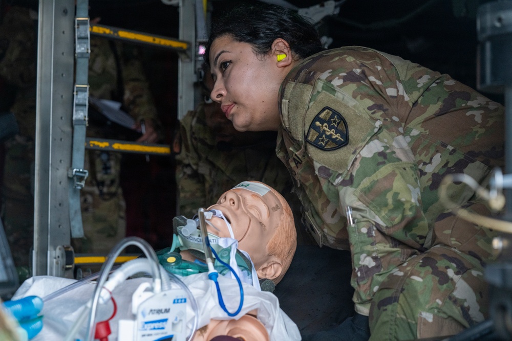 A female military service member in MultiCam/OCP camouflage uniform is conducting medical training using a realistic patient simulation mannequin. The mannequin is equipped with airway management devices including what appears to be an endotracheal tube and oxygen mask. The setting appears to be inside a military aircraft, likely during a medevac or aeromedical evacuation training exercise.