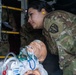 A female military service member in MultiCam/OCP camouflage uniform is conducting medical training using a realistic patient simulation mannequin. The mannequin is equipped with airway management devices including what appears to be an endotracheal tube and oxygen mask. The setting appears to be inside a military aircraft, likely during a medevac or aeromedical evacuation training exercise.