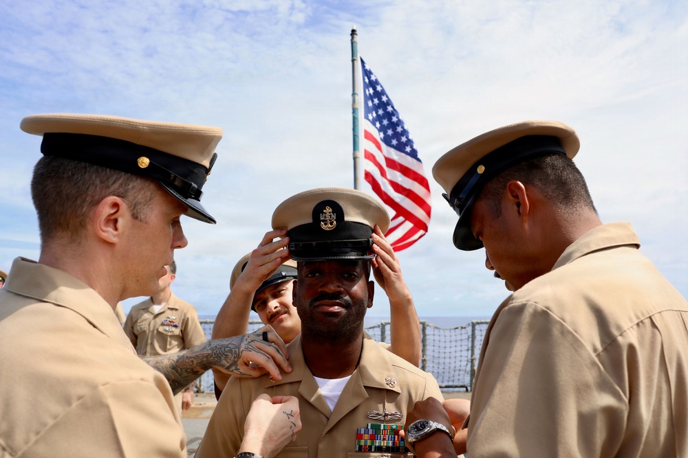 A U.S. Navy promotion or advancement ceremony is taking place on the deck of a naval vessel. The central sailor is having his chief petty officer cover (hat) placed on his head by fellow sailors, a traditional part of the Navy's Chief Petty Officer pinning/advancement ceremony. An American flag is visible in the background, flying from a mast aboard the ship.