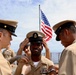 A U.S. Navy promotion or advancement ceremony is taking place on the deck of a naval vessel. The central sailor is having his chief petty officer cover (hat) placed on his head by fellow sailors, a traditional part of the Navy's Chief Petty Officer pinning/advancement ceremony. An American flag is visible in the background, flying from a mast aboard the ship.
