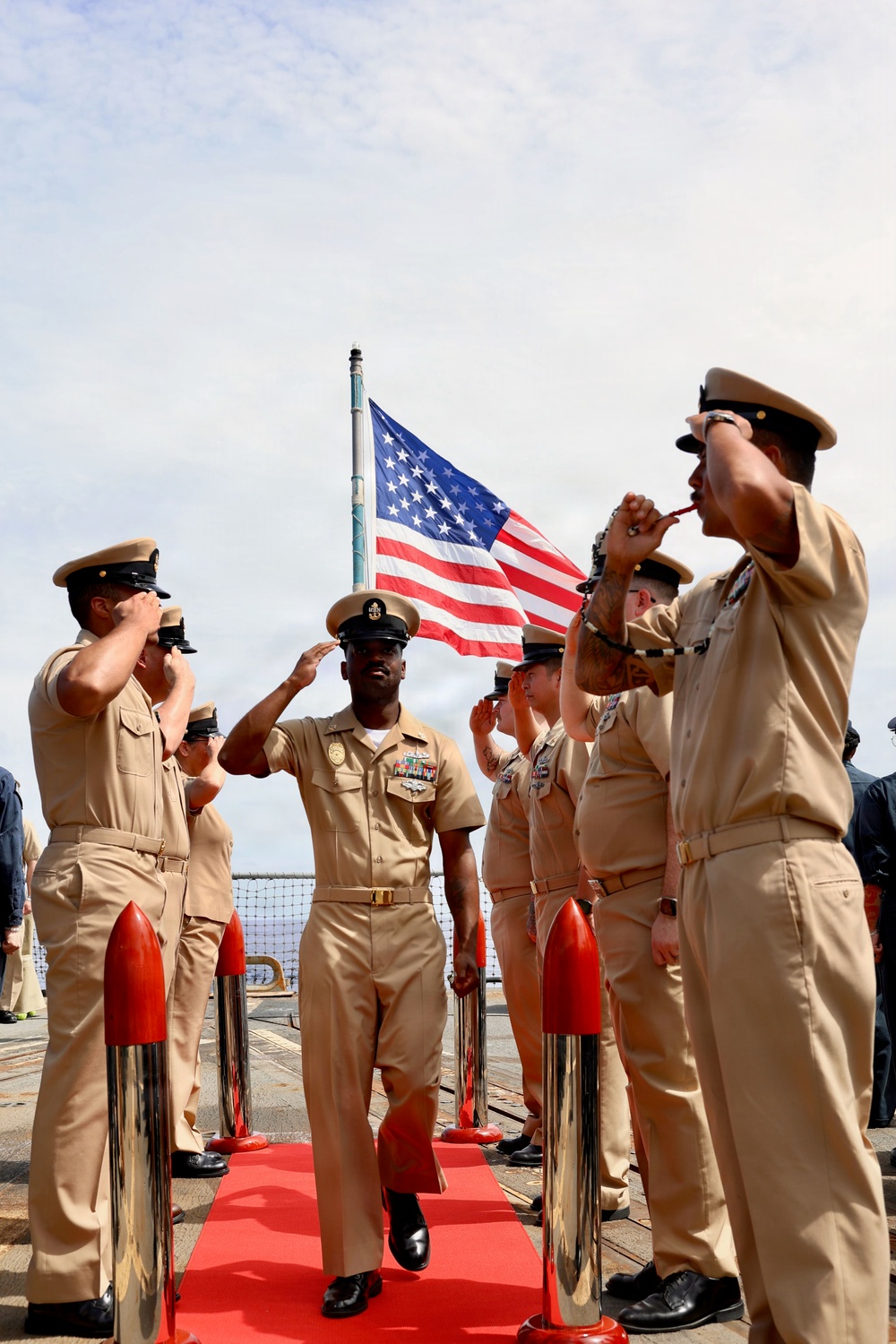 A formal U.S. Navy sideboy ceremony is taking place on what appears to be a ship's deck, with a senior enlisted sailor walking a red carpet while being rendered honors. Personnel on both sides are saluting and piping the side using bosun's whistles, with the American flag flying prominently in the background. The central figure displays multiple service ribbons and a warfare device on their uniform, indicating a senior non-commissioned officer.