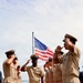 A formal U.S. Navy sideboy ceremony is taking place on what appears to be a ship's deck, with a senior enlisted sailor walking a red carpet while being rendered honors. Personnel on both sides are saluting and piping the side using bosun's whistles, with the American flag flying prominently in the background. The central figure displays multiple service ribbons and a warfare device on their uniform, indicating a senior non-commissioned officer.