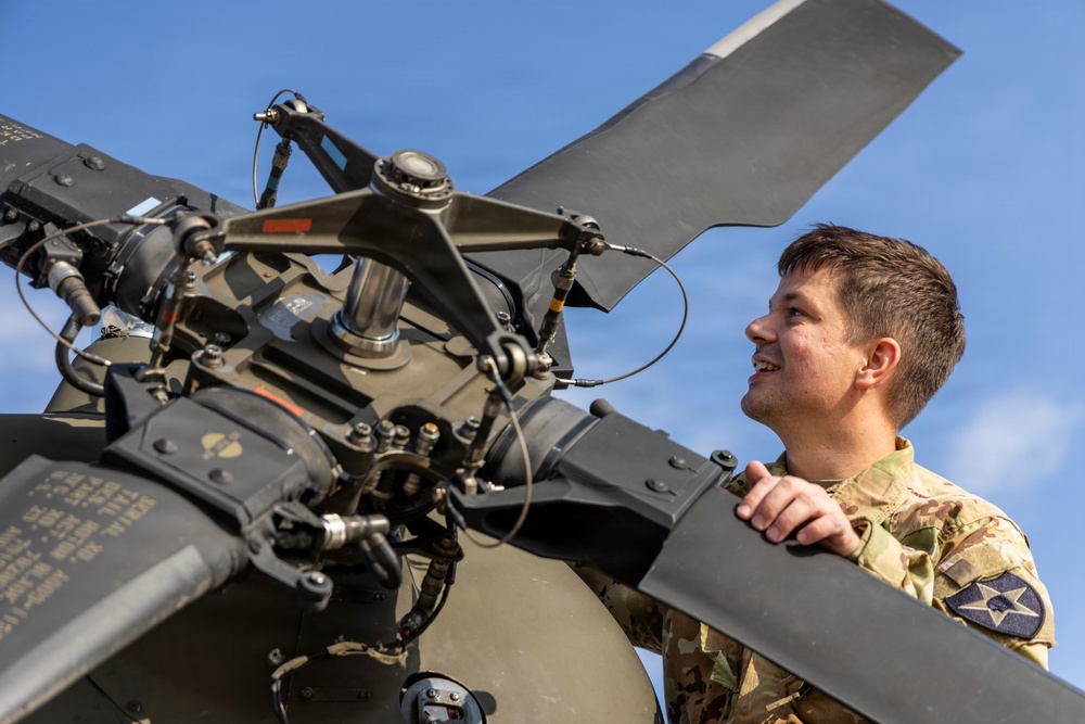 A military service member in camouflage uniform is inspecting or performing maintenance on a helicopter rotor head assembly. The image is taken from a low angle, emphasizing the mechanical complexity of the rotor hub against a blue sky. The individual appears focused and engaged with the aircraft component.