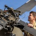 A military service member in camouflage uniform is inspecting or performing maintenance on a helicopter rotor head assembly. The image is taken from a low angle, emphasizing the mechanical complexity of the rotor hub against a blue sky. The individual appears focused and engaged with the aircraft component.