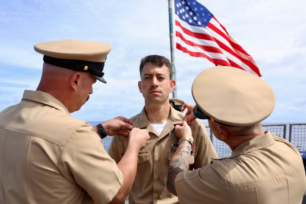 A military promotion or pinning ceremony is taking place outdoors, likely on the deck of a naval vessel. Two senior personnel in U.S. Navy khaki uniforms are pinning insignia or rank devices onto the collar of a third service member standing at attention. An American flag is prominently displayed in the background.