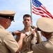 A military promotion or pinning ceremony is taking place outdoors, likely on the deck of a naval vessel. Two senior personnel in U.S. Navy khaki uniforms are pinning insignia or rank devices onto the collar of a third service member standing at attention. An American flag is prominently displayed in the background.