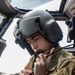 A U.S. Army aviator is seated inside a military helicopter cockpit, wearing a flight helmet with an integrated microphone and appears to be adjusting or connecting communication equipment. The soldier is dressed in OCP (Operational Camouflage Pattern) uniform with a U.S. flag patch visible on the right shoulder and an aviator badge on the chest. The image is a close-up, professionally composed photograph taken inside the aircraft.