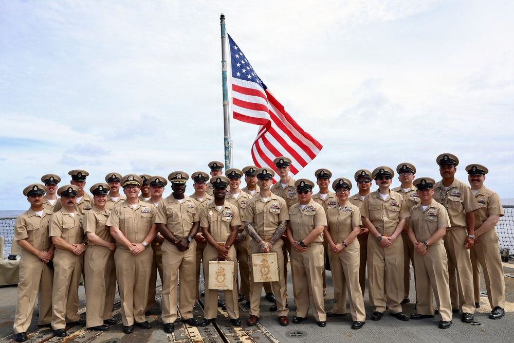 A group of approximately 25 U.S. Navy personnel in khaki service uniforms are posed for a formal group photograph on the deck of a naval vessel. An American flag flies prominently on a flagpole behind the group. Two individuals in the front row are holding what appear to be wooden plaques or awards.