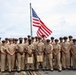 A group of approximately 25 U.S. Navy personnel in khaki service uniforms are posed for a formal group photograph on the deck of a naval vessel. An American flag flies prominently on a flagpole behind the group. Two individuals in the front row are holding what appear to be wooden plaques or awards.