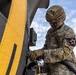 A U.S. Army soldier in MultiCam/OCP camouflage uniform is performing maintenance or fueling operations on a military helicopter with a distinctive yellow and gray paint scheme. The soldier is wearing a combat helmet and tan tactical gloves while handling a hose or cable connected to the aircraft. The image is taken from a low angle against a partly cloudy sky, emphasizing the scale of the aircraft.