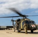 A military medevac helicopter, identified as a UH-60 Black Hawk with red cross markings and the number '023', is running on a tarmac with its rotors spinning. Two ground crew personnel in military combat uniforms and flight helmets are conducting pre-flight or post-flight checks alongside the aircraft. A second helicopter is visible in the background near a hangar.