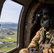 A U.S. Army soldier in full flight gear sits inside a military helicopter, looking out the window at a rural landscape below. The soldier is equipped with a flight helmet, oxygen/communication mask, night vision goggle mount, and tactical vest with a sidearm. The aerial view through the window reveals green agricultural fields, scattered buildings, and distant mountains under a partly cloudy sky.