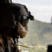 A military aviator or crew member is photographed from inside a helicopter in flight, wearing a tactical flight helmet with integrated communications equipment and dark visor. The visor reflects a cityscape and mountainous terrain below, suggesting the aircraft is flying over an urban area near green hills. The subject appears to be reviewing or handling equipment/documents while airborne.