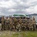 A group of approximately 20 military personnel in U.S. Army OCP (Operational Camouflage Pattern) uniforms pose for a formal group photograph outdoors. They are arranged in two rows — one standing and one kneeling — in front of a white and green van, shipping containers, and a HMMWV (Humvee). The setting appears to be a military installation or forward operating base with mountainous terrain visible in the background.