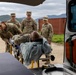 U.S. Army soldiers are loading a patient on a stretcher into the back of an ambulance or medical vehicle. The scene appears to be a medical training exercise or real casualty evacuation taking place in an outdoor military environment. Several soldiers in MultiCam/OCP uniforms are actively participating, with some assisting with the stretcher and others observing.