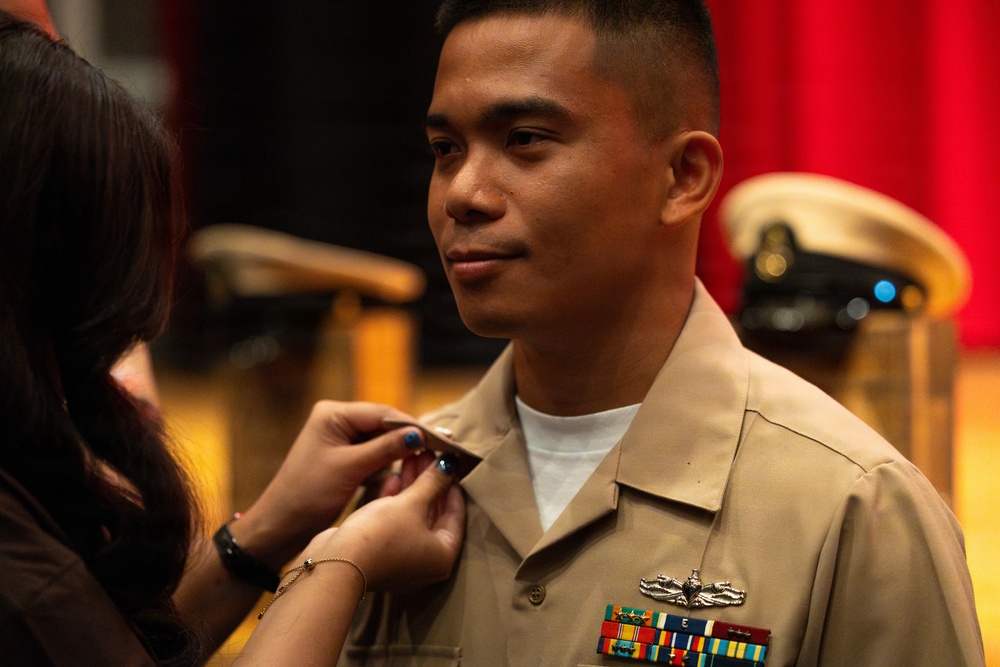 A U.S. Navy service member in a tan/khaki uniform is having a pin or insignia affixed to his collar by another person, likely during a promotion or award ceremony. He is smiling slightly and appears proud. A naval officer's cover (hat) is visible in the blurred background along with red curtains, suggesting a formal ceremonial setting.