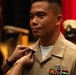 A U.S. Navy service member in a tan/khaki uniform is having a pin or insignia affixed to his collar by another person, likely during a promotion or award ceremony. He is smiling slightly and appears proud. A naval officer's cover (hat) is visible in the blurred background along with red curtains, suggesting a formal ceremonial setting.
