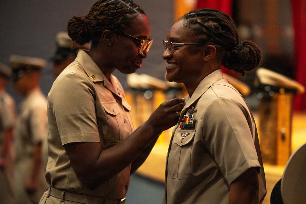 Two female military personnel in tan/khaki uniforms are engaged in what appears to be a pinning ceremony, with one service member affixing an insignia or medal to the other's uniform. Both women are smiling warmly, suggesting a celebratory or promotional occasion. The background shows additional uniformed personnel and what appears to be a formal military ceremony setting.