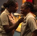 Two female military personnel in tan/khaki uniforms are engaged in what appears to be a pinning ceremony, with one service member affixing an insignia or medal to the other's uniform. Both women are smiling warmly, suggesting a celebratory or promotional occasion. The background shows additional uniformed personnel and what appears to be a formal military ceremony setting.