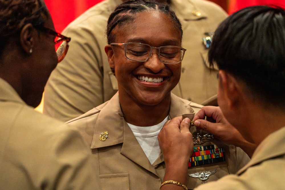 A U.S. Navy service member is being pinned with insignia or a new rank device during what appears to be a military promotion or award ceremony. The central figure is smiling broadly with eyes closed, suggesting a moment of pride and joy. Two other service members in tan Navy uniforms are assisting with the pinning on either side.