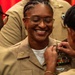 A U.S. Navy service member is being pinned with insignia or a new rank device during what appears to be a military promotion or award ceremony. The central figure is smiling broadly with eyes closed, suggesting a moment of pride and joy. Two other service members in tan Navy uniforms are assisting with the pinning on either side.