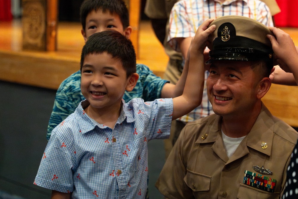 A U.S. Navy Chief Petty Officer in khaki dress uniform is having his cover (hat) placed on his head by children during what appears to be a military promotion or pinning ceremony. The sailor and a young boy in the foreground are both smiling broadly, suggesting a joyful family moment. The setting appears to be an indoor military ceremony hall.