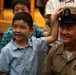 A U.S. Navy Chief Petty Officer in khaki dress uniform is having his cover (hat) placed on his head by children during what appears to be a military promotion or pinning ceremony. The sailor and a young boy in the foreground are both smiling broadly, suggesting a joyful family moment. The setting appears to be an indoor military ceremony hall.