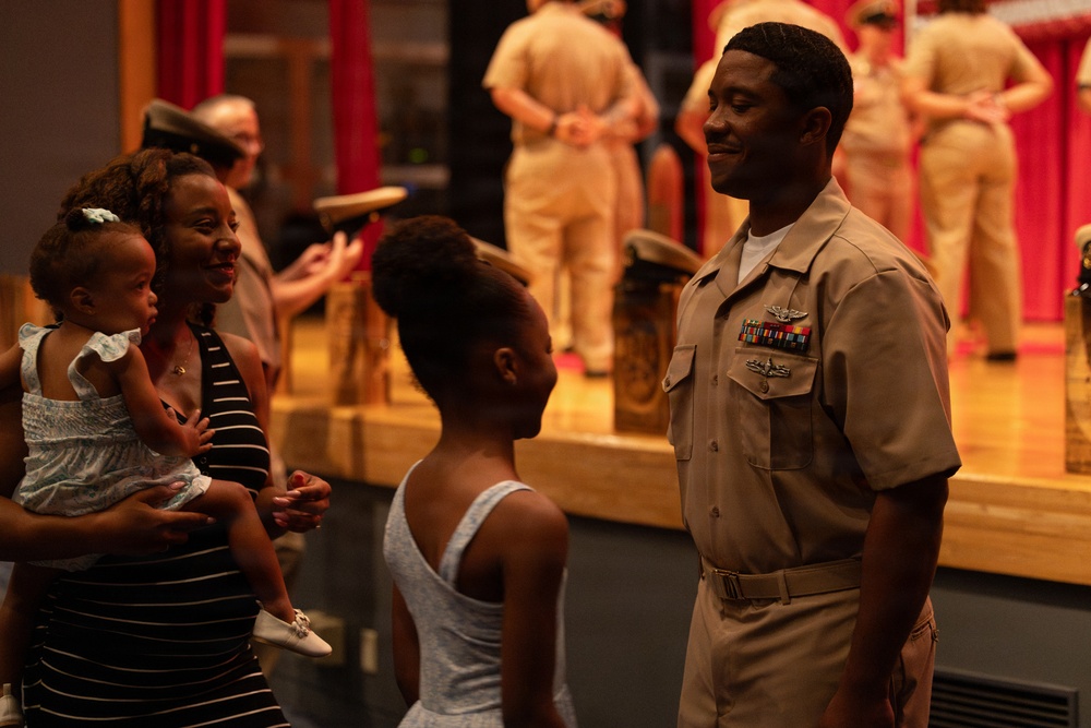A U.S. Navy officer in service khaki uniform is greeting his family, including a woman holding a toddler and a young girl, following what appears to be a military ceremony. The officer is wearing ribbons, a warfare device badge, and rank insignia. In the background, other uniformed personnel are visible on or near a stage with red curtains.