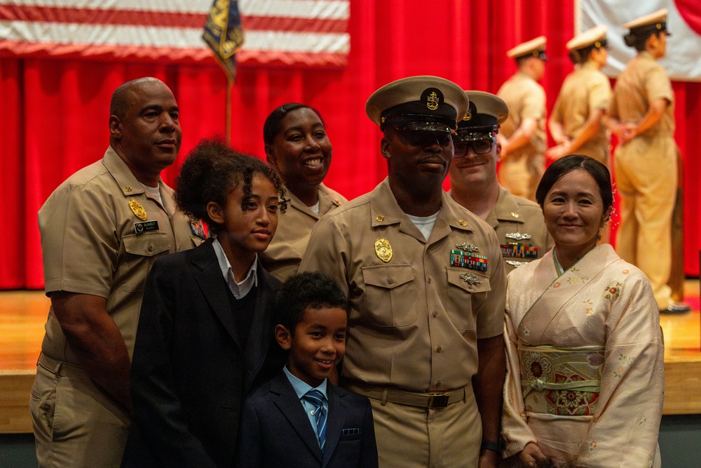 A U.S. Navy promotion or advancement ceremony is taking place in a formal venue decorated with red curtains and American flags. A senior Navy chief petty officer in dress khaki uniform poses with what appears to be his family, including two children and a woman wearing a traditional Japanese kimono. Additional Navy personnel in uniform are visible both in the foreground and on a stage in the background.