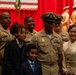 A U.S. Navy promotion or advancement ceremony is taking place in a formal venue decorated with red curtains and American flags. A senior Navy chief petty officer in dress khaki uniform poses with what appears to be his family, including two children and a woman wearing a traditional Japanese kimono. Additional Navy personnel in uniform are visible both in the foreground and on a stage in the background.