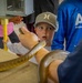 A young boy wearing a gray Hurley cap reacts with wide-eyed surprise as a large snake is brought close to his face during what appears to be an educational animal demonstration. A student wearing a blue 'AVID - Andersen Middle School' shirt stands nearby observing. The setting appears to be an indoor educational or zoo-type event with multiple children present.