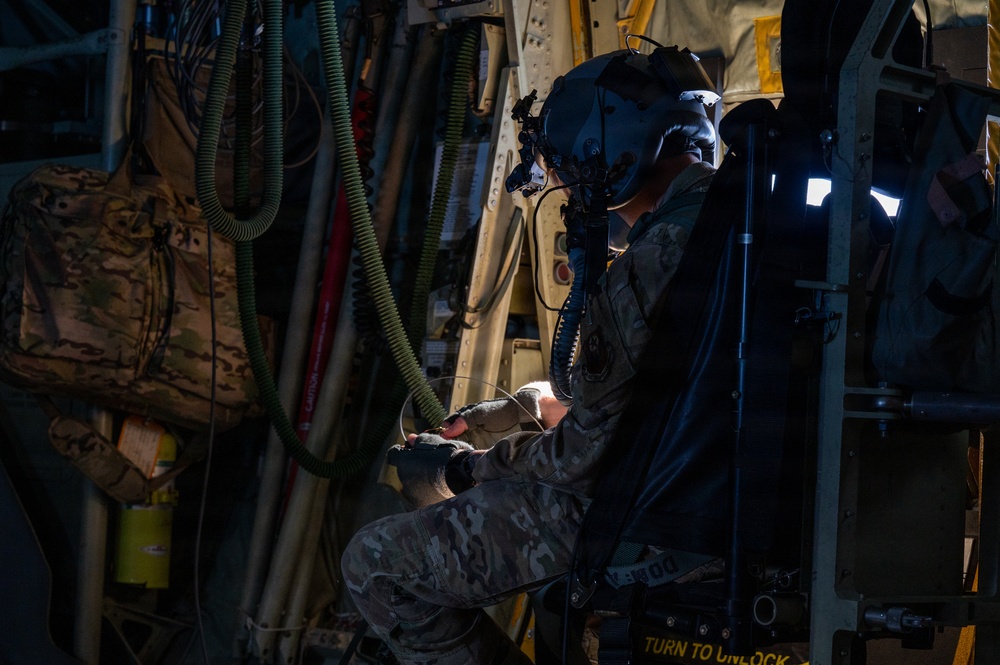 A military aircrew member in full flight gear, including a helmet with night vision goggle mount, is working inside what appears to be a large military transport aircraft. The individual is wearing multicam/OCP camouflage uniform and tactical gloves while manipulating equipment near a door or hatch mechanism. The interior environment is dimly lit, revealing various hoses, cables, equipment bags, and aircraft structural components.