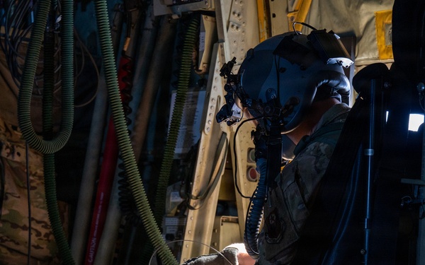 A military aircrew member in full flight gear, including a helmet with night vision goggle mount, is working inside what appears to be a large military transport aircraft. The individual is wearing multicam/OCP camouflage uniform and tactical gloves while manipulating equipment near a door or hatch mechanism. The interior environment is dimly lit, revealing various hoses, cables, equipment bags, and aircraft structural components.