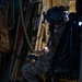 A military aircrew member in full flight gear, including a helmet with night vision goggle mount, is working inside what appears to be a large military transport aircraft. The individual is wearing multicam/OCP camouflage uniform and tactical gloves while manipulating equipment near a door or hatch mechanism. The interior environment is dimly lit, revealing various hoses, cables, equipment bags, and aircraft structural components.