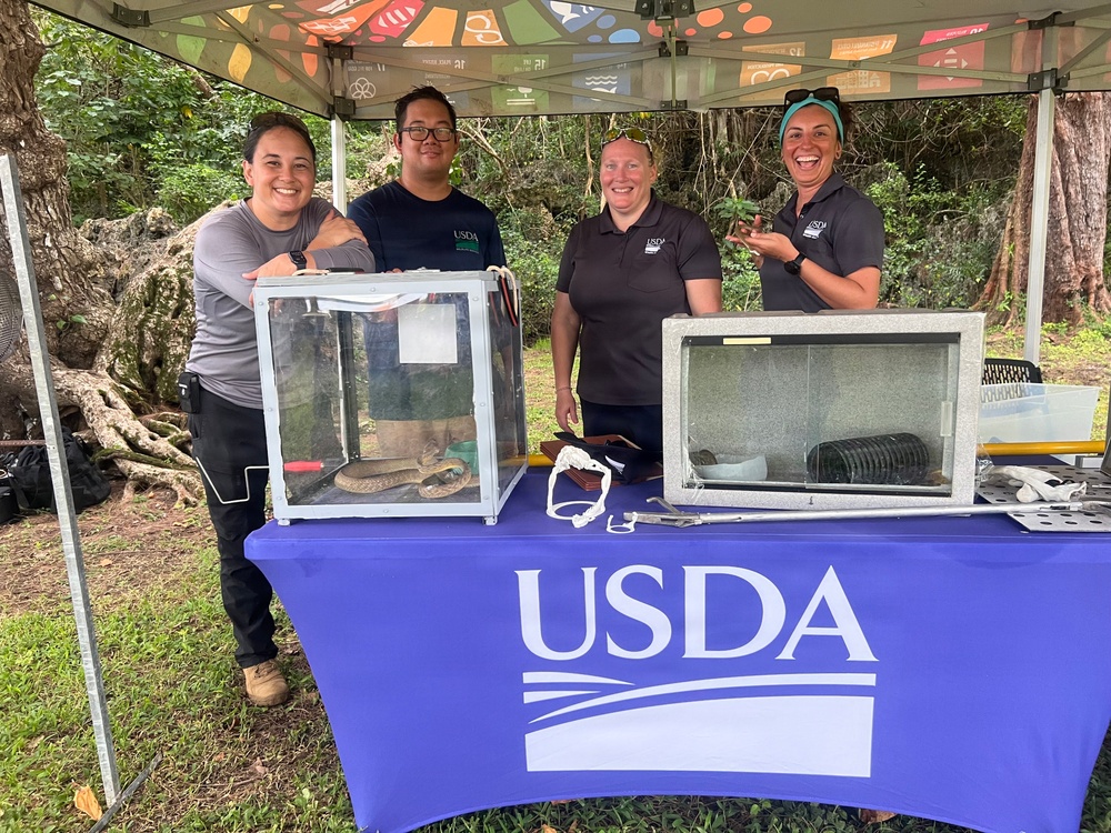 Four USDA personnel are standing behind a table draped with a blue USDA-branded tablecloth at an outdoor educational or outreach event. Two glass display enclosures are on the table, with one visibly containing what appears to be a brown tree snake. The setting is a forested outdoor area under a colorful canopy tent.