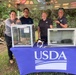 Four USDA personnel are standing behind a table draped with a blue USDA-branded tablecloth at an outdoor educational or outreach event. Two glass display enclosures are on the table, with one visibly containing what appears to be a brown tree snake. The setting is a forested outdoor area under a colorful canopy tent.