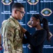 A woman in a black long-sleeve shirt is pinning something onto the chest of a male service member wearing OCP (Operational Camouflage Pattern) military uniform. The backdrop displays the 'Andersen Fire Department' logo repeatedly, suggesting this is an official military or fire department ceremony. The act of pinning is consistent with a promotion, award, or badge ceremony.