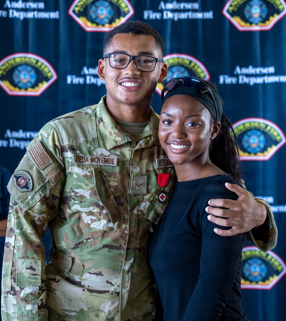 A U.S. Air Force servicemember named Beda Moyembe, wearing OCP (Operational Camouflage Pattern) uniform with a red cross medal pinned to his chest, poses with a young woman in front of an Andersen Fire Department backdrop. The servicemember appears to have received a medical/lifesaving award or commendation. Both individuals are smiling and appear to be celebrating the recognition.