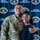 A U.S. Air Force servicemember named Beda Moyembe, wearing OCP (Operational Camouflage Pattern) uniform with a red cross medal pinned to his chest, poses with a young woman in front of an Andersen Fire Department backdrop. The servicemember appears to have received a medical/lifesaving award or commendation. Both individuals are smiling and appear to be celebrating the recognition.