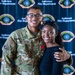 A U.S. Air Force servicemember named Beda Moyembe, wearing OCP (Operational Camouflage Pattern) uniform with a red cross medal pinned to his chest, poses with a young woman in front of an Andersen Fire Department backdrop. The servicemember appears to have received a medical/lifesaving award or commendation. Both individuals are smiling and appear to be celebrating the recognition.